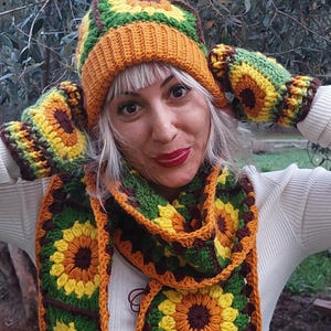 Puede incluir: Un conjunto de gorro, bufanda y guantes de punto de ganchillo con forma de girasol en tonos marrones, verdes, amarillos y naranjas. El gorro tiene un pompón marrón en la parte superior. La bufanda tiene flecos en los extremos.