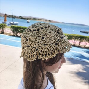 May include: A beige crochet bucket hat featuring an intricate openwork pattern with a scalloped edge. The hat is constructed from a light-colored yarn and displays a detailed, repeating circular design throughout the crown and the brim.