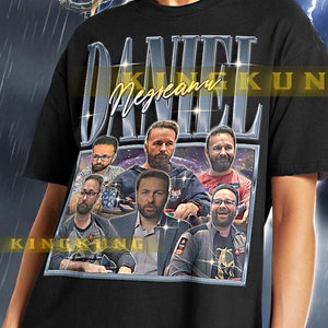 Pode incluir: Camiseta unisex preta com uma estampa gráfica de um grupo de pessoas e o texto "DANIEL NEGREANU KING". A camiseta está sendo modelada por uma pessoa vestindo calças pretas.