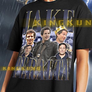 May include: Black unisex t-shirt featuring a graphic print of a group of people and the text "Jake Peralta" in gold lettering.