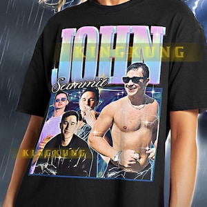 May include: Black unisex t-shirt featuring a retro-style graphic with the name "JOHN" in a colorful, stylized font. The graphic includes images of a man and the name "Scanmit". The shirt is worn by a person.