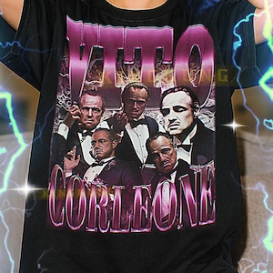 May include: Black t-shirt featuring a graphic of the word "VITO" in a purple gradient font above a collage of men in suits. The word "CORLEONE" is below in a similar font. The shirt is unisex.