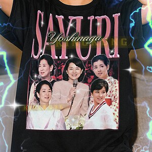 Könnte beinhalten: Schwarzes Unisex-T-Shirt mit einem rosa "Sayuri Yoshinaga"-Aufdruck und einem Foto von fünf Frauen. Das T-Shirt hat einen Rundhalsausschnitt und kurze Ärmel. Der Text am unteren Rand des T-Shirts ist nicht enthalten.
