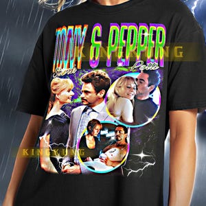 May include: Black unisex t-shirt featuring a colorful graphic of the words "Tony & Pepper Potts" with images of the characters. The shirt has a vintage, retro style.