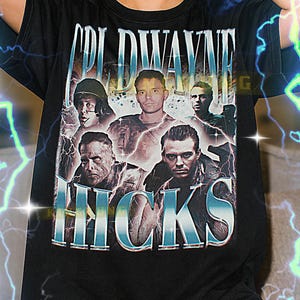 Puede incluir: Camiseta negra con un diseño gráfico con el texto "COLD WAYNE HICKS" e imágenes de varios hombres. La camiseta la lleva una persona con el pelo corto y rubio. La camiseta es unisex.