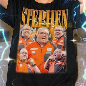 May include: Black t-shirt featuring a graphic of Stephen Bunting, a darts player, in an orange and black uniform. The shirt has the name "Stephen Bunting" in gold lettering. The model is wearing an XL shirt.
