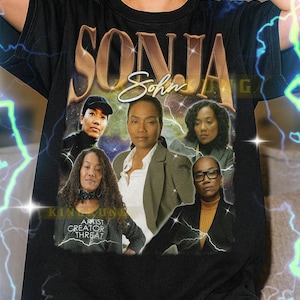 May include: Black unisex t-shirt featuring a graphic of Sonja Sohn and other women. The shirt has the text "SONJA" in gold lettering. The text "ARTIST CREATOR THREAT" is also visible. The model is wearing an XL shirt.