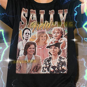May include: Black unisex t-shirt featuring a vintage-style graphic of Sally Field with the text "SALLY Field" in gold lettering. The shirt has a crew neck and short sleeves. The graphic includes several images of Sally Field. The shirt is made of a soft material.