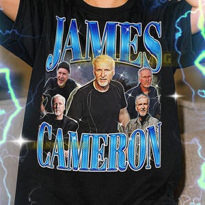 May include: Black t-shirt featuring the name "JAMES CAMERON" in blue, stylized text. The shirt includes a graphic of James Cameron and other men. The shirt is a unisex design.