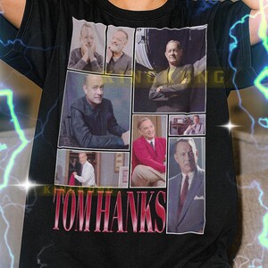 Op de afbeelding: Zwart T-shirt met een collage van Tom Hanks-afbeeldingen en de tekst "TOM HANKS" in roze en rood. Dit unisex shirt is een casual stijl.