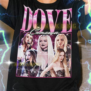 Op de afbeelding: Zwart T-shirt met een roze afbeelding met de tekst "DOVE Cameron" en een foto van Dove Cameron. Het shirt heeft een ronde hals en korte mouwen. De tekst onderaan het shirt is niet inbegrepen.