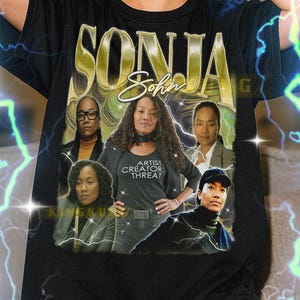 May include: Black unisex t-shirt featuring the name "SONJA" in gold lettering above a graphic of five women. One woman wears a shirt that says "ARTIST CREATOR THREAT". The shirt fits true to size.