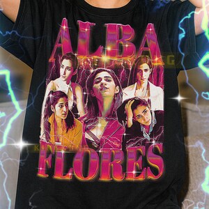 以下が含まれることがあります： ピンクとオレンジ色の「ALBA FLORES」の文字が入ったグラフィックデザインの黒いTシャツ。デザインには、稲妻の効果を持つ女性の画像が含まれています。このTシャツはユニセックスです。
