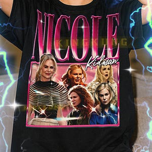 May include: Black t-shirt featuring the name "NICOLE" in pink, with a pink framed image of Nicole Kidman. The shirt has a vintage style design with a retro aesthetic.