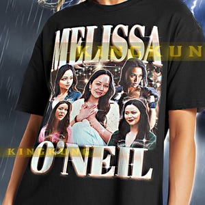 Puede incluir: Camiseta negra con un estampado gráfico de Melissa O'Neil y el texto "MELISSA O'NEIL" en una fuente brillante. La modelo lleva una camiseta talla XL.