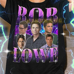 May include: Black t-shirt featuring the name "ROB LOWE" in large, purple, glittery letters. The shirt has a collage of Rob Lowe's images. The text "ROB" is arched above the images. The text at the bottom of the shirt is "LOWE".