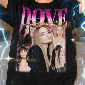 May include: Black unisex t-shirt featuring a graphic of a woman with the text "DOVE" in pink. The shirt includes multiple images of the woman. The text "KIMOKUNG" is also visible.