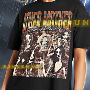 以下が含まれることがあります： 前面に「Other Mother The Belladam」というテキストと、カートゥーンキャラクターのグループのグラフィックプリントが施された黒いTシャツです。