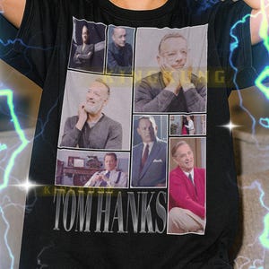 May include: Black t-shirt featuring a collage of images of Tom Hanks with the text "TOM HANKS" in a bold, silver font. The shirt is a unisex style.