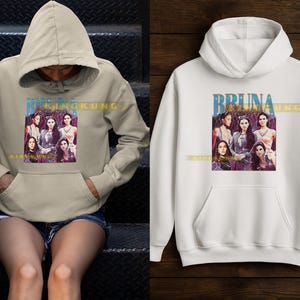 May include: Beige and white hooded sweatshirts. Each sweatshirt features a graphic of a group of women with the text "BRUNA KING KUNG" in a blue and gold font. The sweatshirts have a front pocket and a drawstring hood.