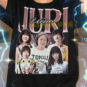 Può includere: T-shirt unisex nera con una grafica di un gruppo di donne e la parola "JURI". La maglietta ha anche le parole "TONGA" e "FORD TATE PARK". La modella indossa una maglietta XL.