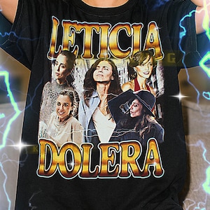 May include: Black unisex t-shirt with the gold text "LETICIA DOLERA" above a collage of women. The shirt has a crew neck and is a classic style.