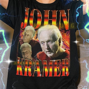 Könnte beinhalten: Schwarzes T-Shirt mit einer Grafik von John Kramer in Rot und Orange. Der Text "JOHN KRAMER" ist in einer kräftigen, stilisierten Schriftart dargestellt. Das Shirt ist ein Unisex-Stil.