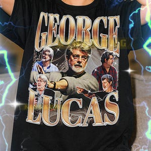 May include: Black t-shirt featuring the name "GEORGE LUCAS" in gold lettering with a vintage design. The shirt includes a collage of images of George Lucas. The text at the bottom of the shirt is not included.