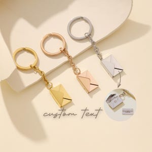 May include: Three metal keyrings in gold, rose gold, and silver, each with a small envelope charm. The keyrings are connected to a ring and chain. The words "custom text" are written in cursive.