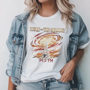 May include: White t-shirt featuring a graphic of a radio tower with a swirling orange and yellow design. The text "WSQK - THE SQUAWK" and "94.5 FM" are printed above and below the graphic.