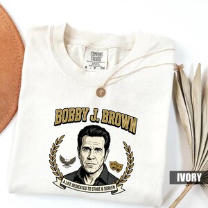 May include: Ivory-colored t-shirt featuring a portrait of Bobby J. Brown, framed by a laurel wreath, an eagle, and theater masks. The text "BOBBY J. BROWN" is in gold lettering above the portrait, with "A LIFE DEDICATED TO STAGE & SCREEN" below.