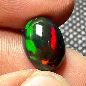 May include: A polished, oval-shaped black opal gemstone. The opal displays vibrant flashes of green, red, and orange colors within its dark surface. The gemstone is held between fingers, showcasing its reflective qualities.