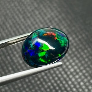 May include: A polished, oval-shaped black opal gemstone with vibrant flashes of green, blue, and red. The gemstone is held by a pair of silver tweezers, showcasing its iridescent play-of-color. The opal is a natural gemstone.