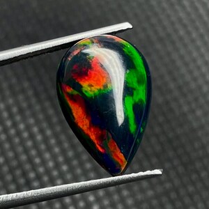 May include: A teardrop-shaped black opal gemstone with vibrant internal colors of red, green, and yellow. The polished stone is held by silver tweezers against a dark gray background. The opal's surface reflects light.