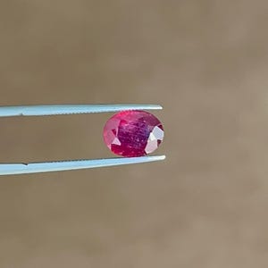 May include: A faceted oval-shaped ruby gemstone with a deep red hue and visible internal inclusions, held securely between the two thin, light-colored metal tips of a pair of precision tweezers, showcasing its polished surface and clarity.