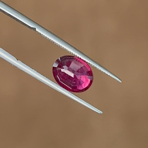 Può includere: Una gemma di rubino dal taglio ovale vibrante con una tonalità rosso rosato intenso e una superficie sfaccettata, tenuta saldamente da una pinzetta in metallo color argento. La gemma mostra chiarezza interna e riflessi di luce.