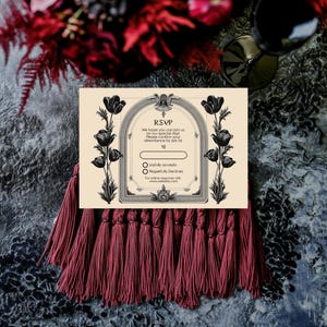 May include: An RSVP card with a cream background, black floral illustrations, and the text "RSVP". The card is decorated with black tulips and framed by an ornate border. Below the card is a burgundy tassel.