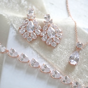 May include: A rose gold bridal jewelry set with a necklace, bracelet, and earrings. The set features clear cubic zirconia stones in a teardrop and round shape.