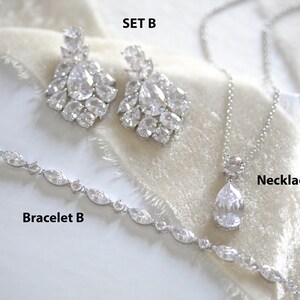 May include: A set of bridal jewelry featuring a silver necklace with a teardrop-shaped crystal pendant, a silver bracelet with oval-shaped crystals, and a pair of silver earrings with large, multi-faceted crystals. The set is labeled "Set B", "Necklace A", and "Bracelet B".