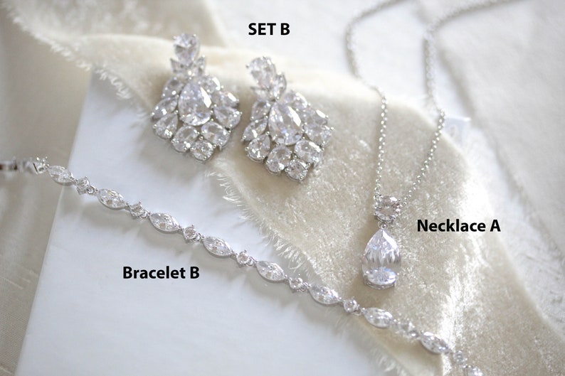 May include: A set of bridal jewelry featuring a silver necklace with a teardrop-shaped crystal pendant, a silver bracelet with oval-shaped crystals, and a pair of silver earrings with large, multi-faceted crystals. The set is labeled "Set B", "Necklace A", and "Bracelet B".