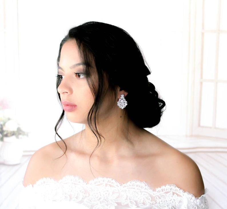 May include: A woman with long black hair wearing a white off-the-shoulder lace dress and sparkling crystal earrings.