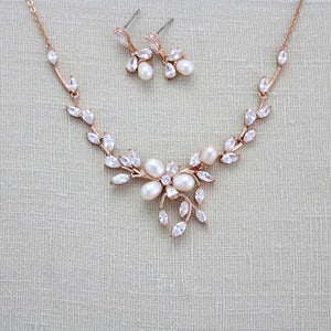 May include: A rose gold necklace and earrings set with a floral design and clear crystals. The necklace features a delicate chain with a pendant that resembles a flower with leaves. The earrings are small and dangle with a single pearl.