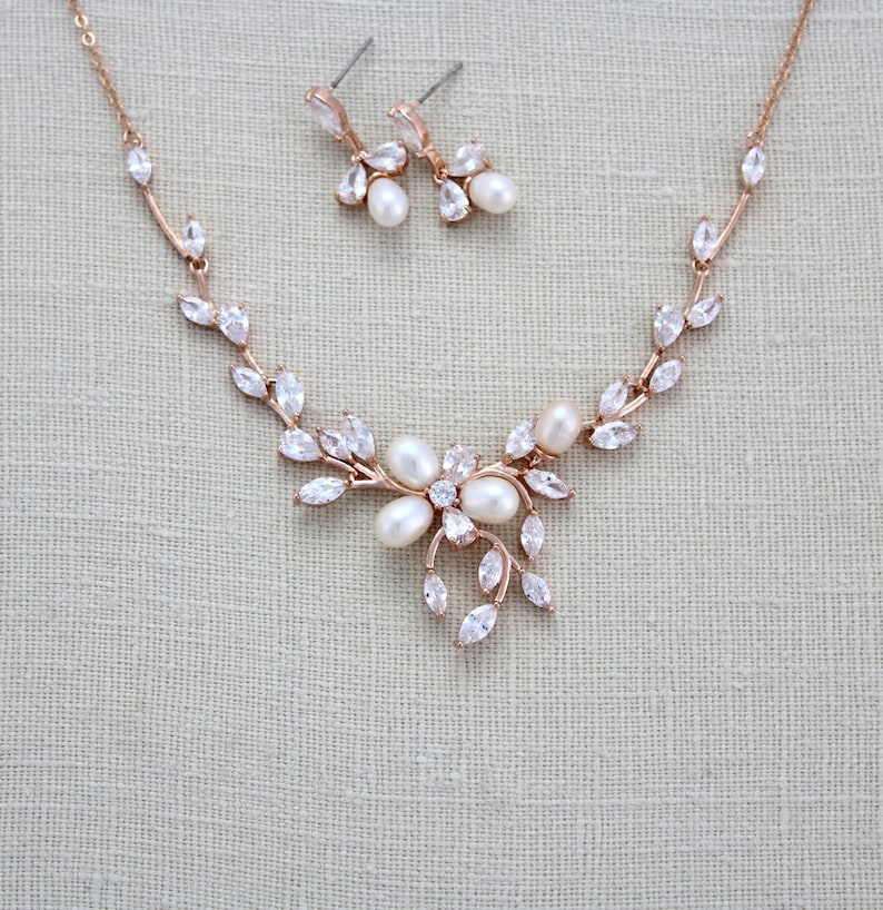 May include: A rose gold necklace and earrings set with a floral design and clear crystals. The necklace features a delicate chain with a pendant that resembles a flower with leaves. The earrings are small and dangle with a single pearl.