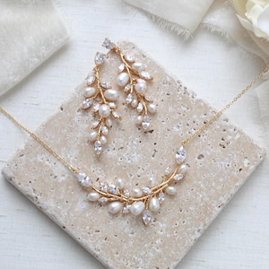 May include: A gold-coloured jewellery set comprising a necklace and matching earrings. The necklace and earrings are decorated with white pearls and clear, sparkling crystals, arranged in a delicate, leaf-like design. The set is displayed on a textured, light-coloured surface.