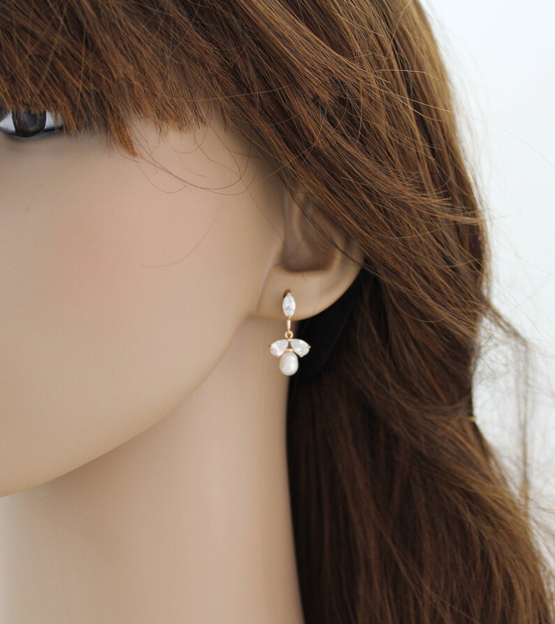 May include: Gold dangle earrings with a pearl and clear stones. The earrings are on a mannequin's ear.