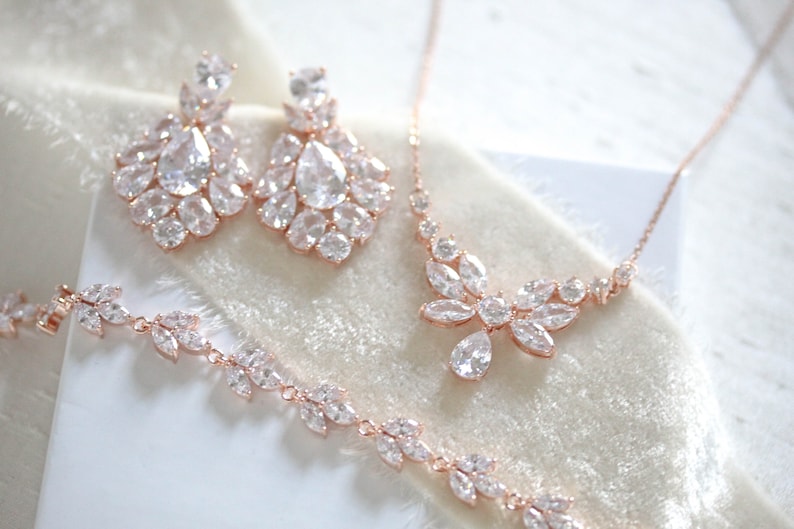 May include: A rose gold bridal jewelry set with a necklace, earrings, and bracelet. The set features a delicate floral design with sparkling clear stones.