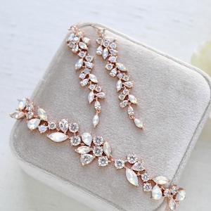 May include: A rose gold jewellery set comprising a bracelet and earrings. The set is decorated with clear and opalescent crystals in a floral pattern. The jewellery is presented on a grey velvet surface.