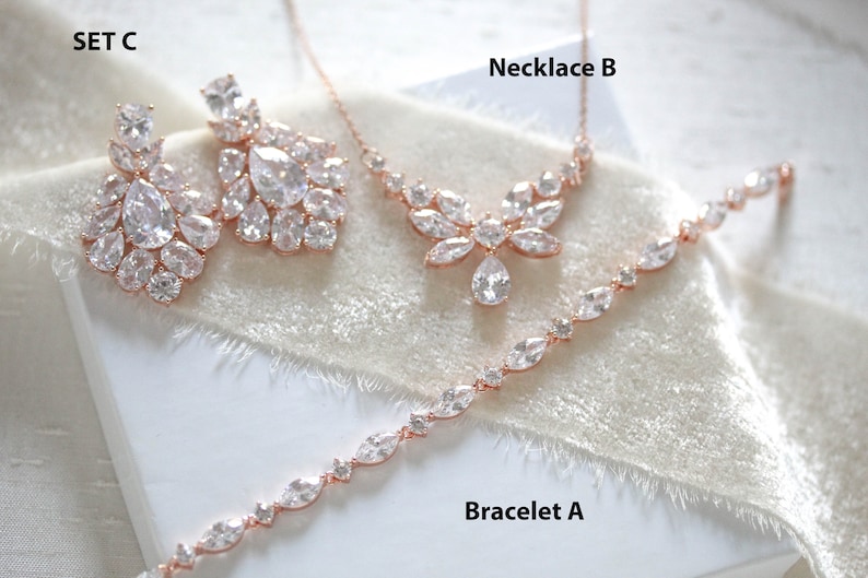 May include: A set of rose gold jewelry with clear cubic zirconia stones. The set includes a necklace, bracelet, and earrings. The necklace is labeled "Necklace B", the bracelet is labeled "Bracelet A", and the earrings are labeled "SET C".