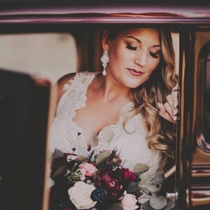 Puede incluir: Una novia con un vestido de novia de encaje blanco con escote pronunciado, sosteniendo un ramo de flores burdeos, rosas y blancas. Lleva pendientes colgantes de cristal y tiene el pelo ondulado. La foto está tomada dentro de un coche antiguo.