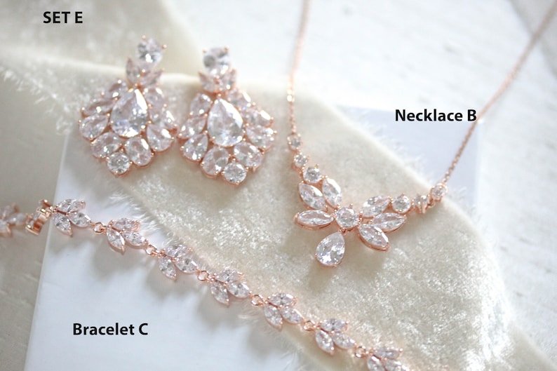 May include: A rose gold bridal jewelry set with a necklace, earrings, and bracelet. The set features a delicate floral design with sparkling clear stones. The necklace is labeled "Necklace B", the earrings are labeled "SET E", and the bracelet is labeled "Bracelet C".